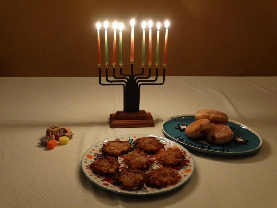 A menorah is lit, framed by the symbols of Hanukkah: Dreidels, Latkes, and Sufganiyot. 