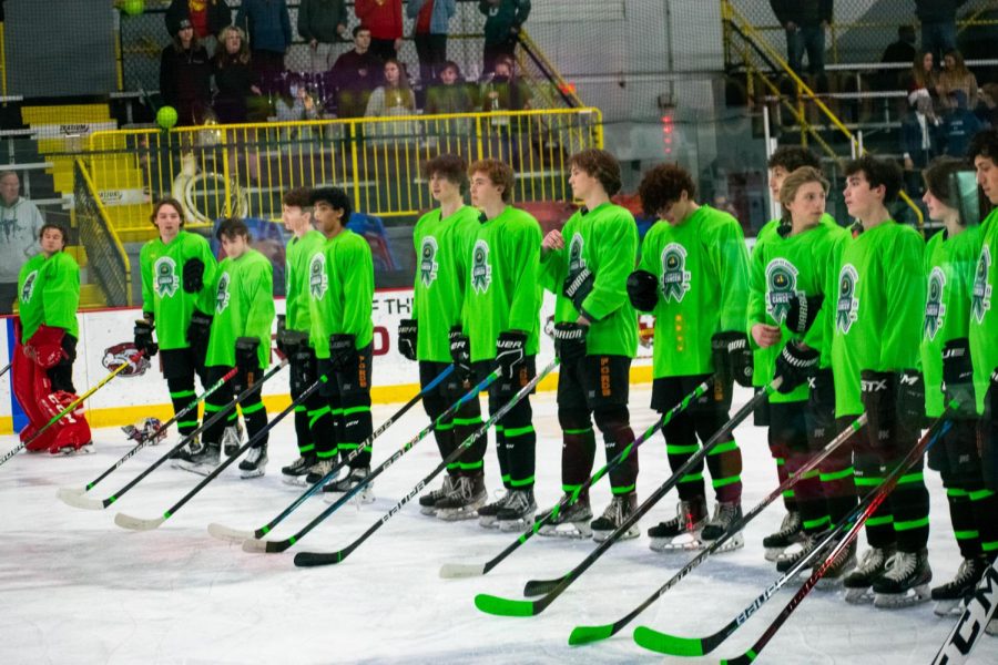 The Varsity Ice Hockey team lines up for the national anthem, sporting green jerseys, stick tape, and helmet decals. 